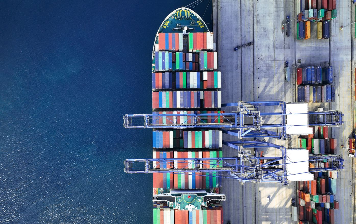 Direct overhead view of a container ship docked at a terminal, with gantry cranes unloading colorful shipping containers beside deep blue water.