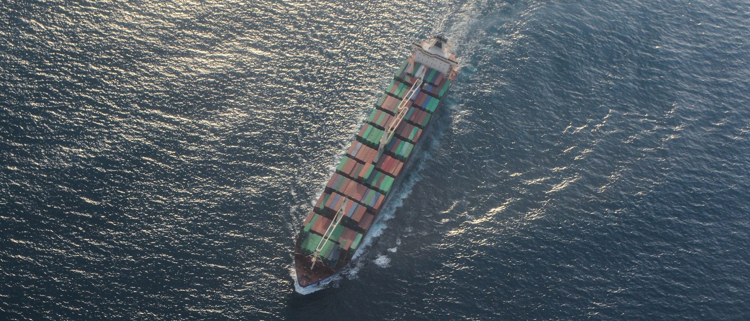 Aerial view of a fully laden container ship underway at sea, leaving a long white wake across dark blue water.